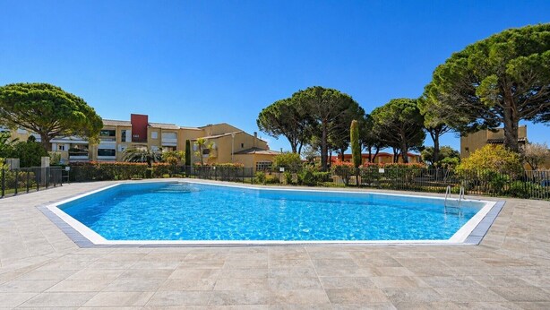 Una piscina rettangolare con bordo piastrellato, circondata da alberi e edifici.