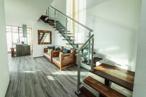 A spacious living area with a wooden sofa, a glass staircase, and a kitchen in the background.