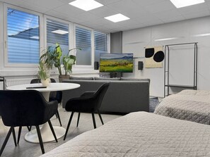 Ein modernes Hotelzimmer mit Bett, Schreibtisch mit Stuhl, Fernseher und Fenster mit Jalousien.