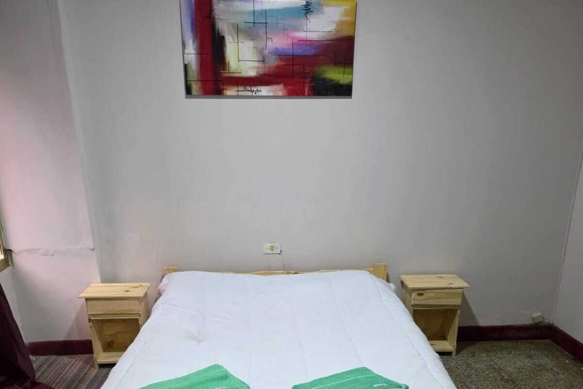 Quarto de hotel com uma cama, duas mesinhas de cabeceira de madeira, um quadro na parede e um tapete listrado.