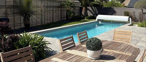 Uma mesa de madeira à beira da piscina com cadeiras, uma piscina e uma cerca de madeira ao fundo.