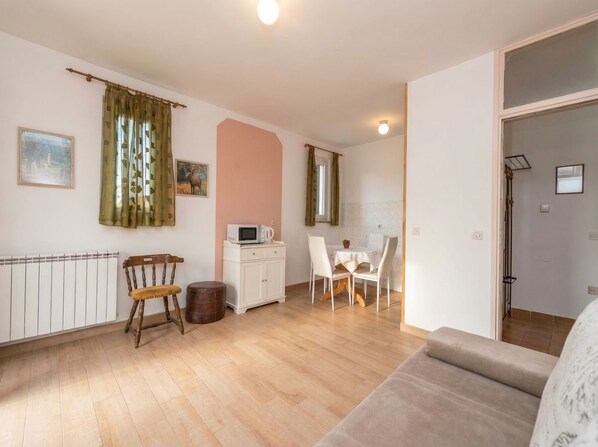 Una stanza con pavimento in legno, un termosifone bianco, una sedia di legno, un armadietto bianco con un forno a microonde, un tavolo con sedie e un divano.