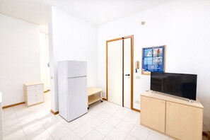 Habitació petita i lluminosa amb un frigorífic blanc, un televisor sobre un moble de fusta, una porta amb mirall i un cartell a la paret.