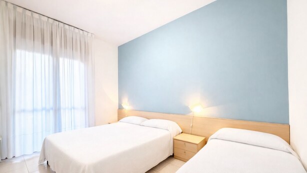 A hotel room with two beds, a nightstand, a light blue wall, and a window with curtains. - Villa Luisa 2 - Carraro Immobiliare Jesolo (Lido di Jesolo)