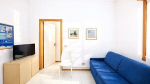A living room with a blue sofa, a white chair, a TV on a wooden cabinet, and two framed pictures on the wall. - Villa Luisa 2 - Carraro Immobiliare Jesolo (Lido di Jesolo)