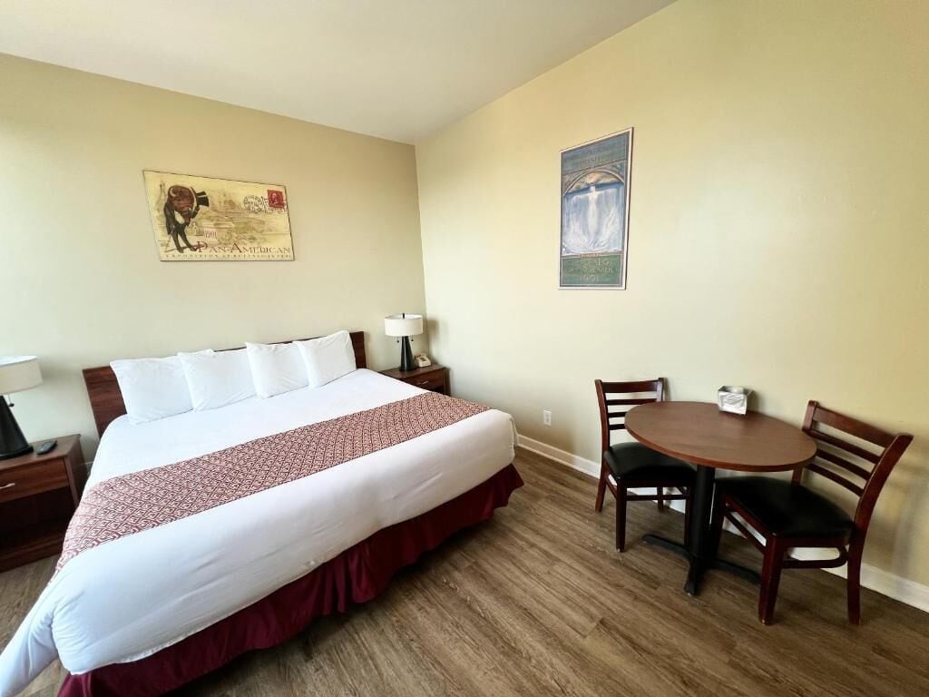 Una habitación de hotel con una cama grande, una mesita pequeña, dos sillas, una mesita de noche y dos cuadros colgados en la pared.