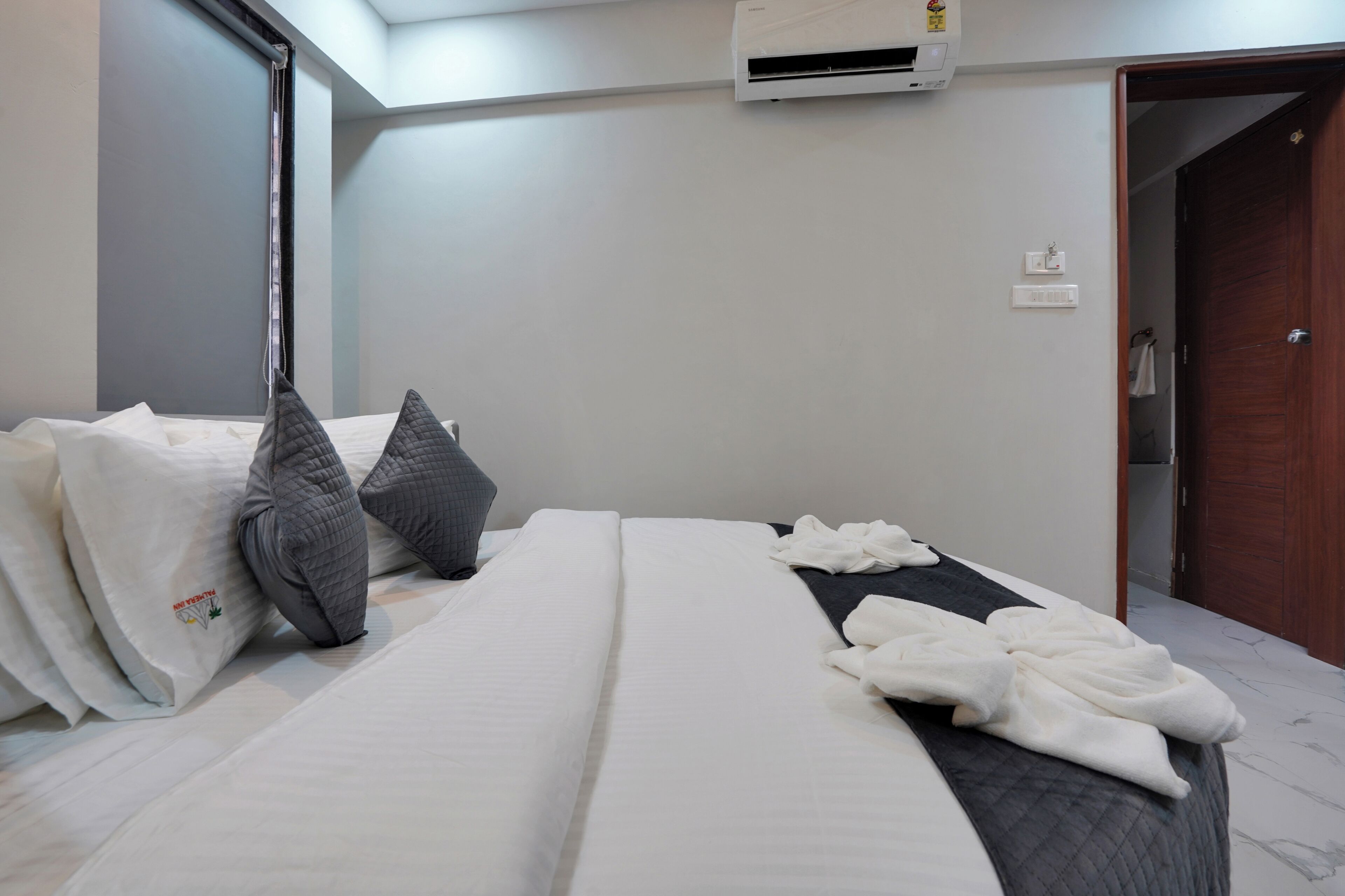Habitación de hotel con cama, almohadas, una manta, unidad de aire acondicionado, puerta y un perchero para toallas.