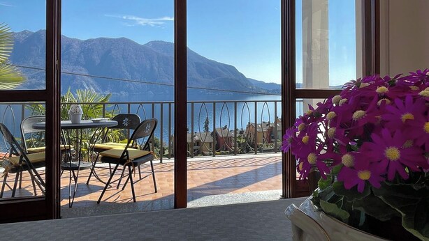 A balcony with a table and chairs, a potted plant, and a view of a lake and mountains.