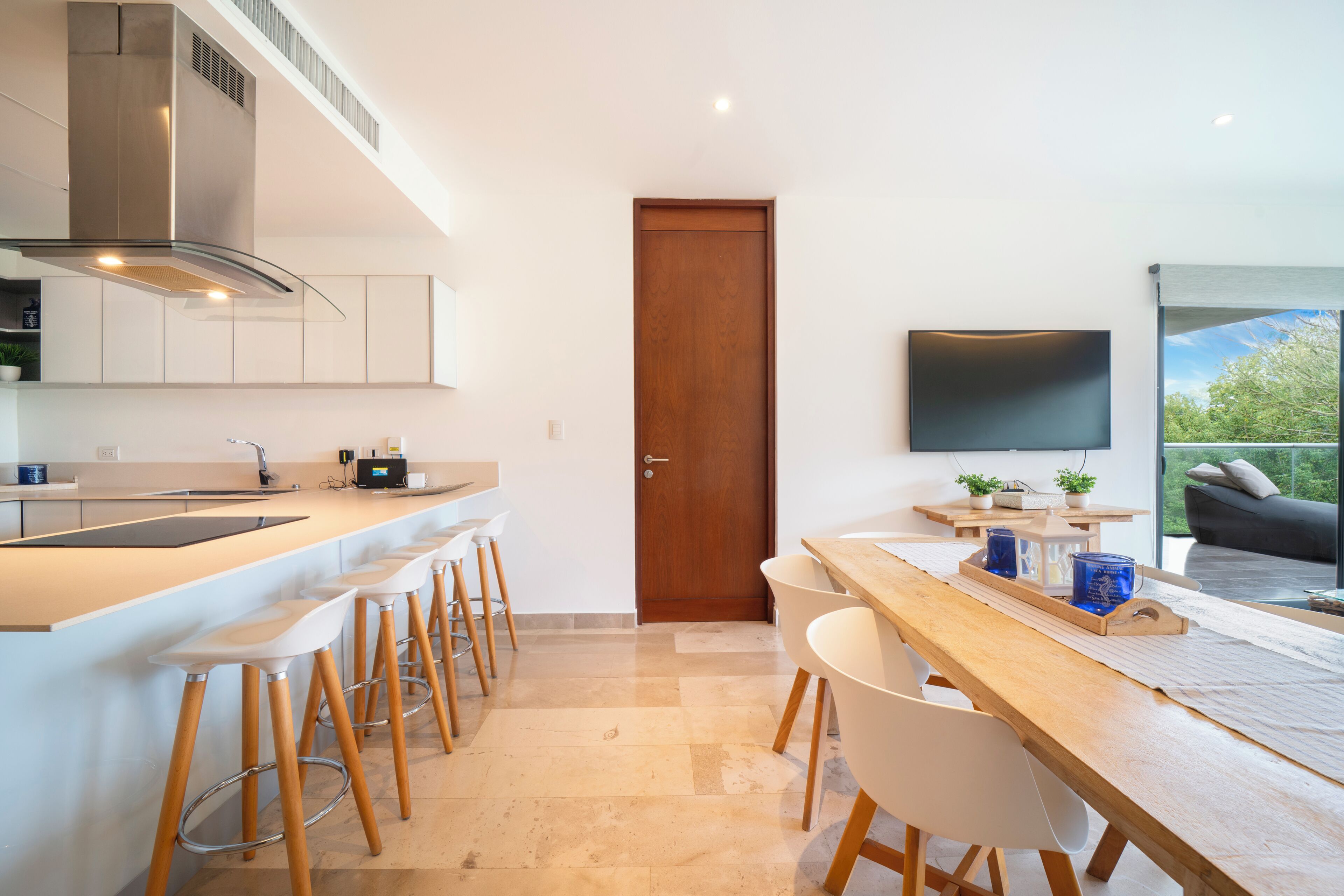 A modern kitchen with a central island, bar stools, a dining table with chairs, a wooden door, and a TV mounted on the wall.