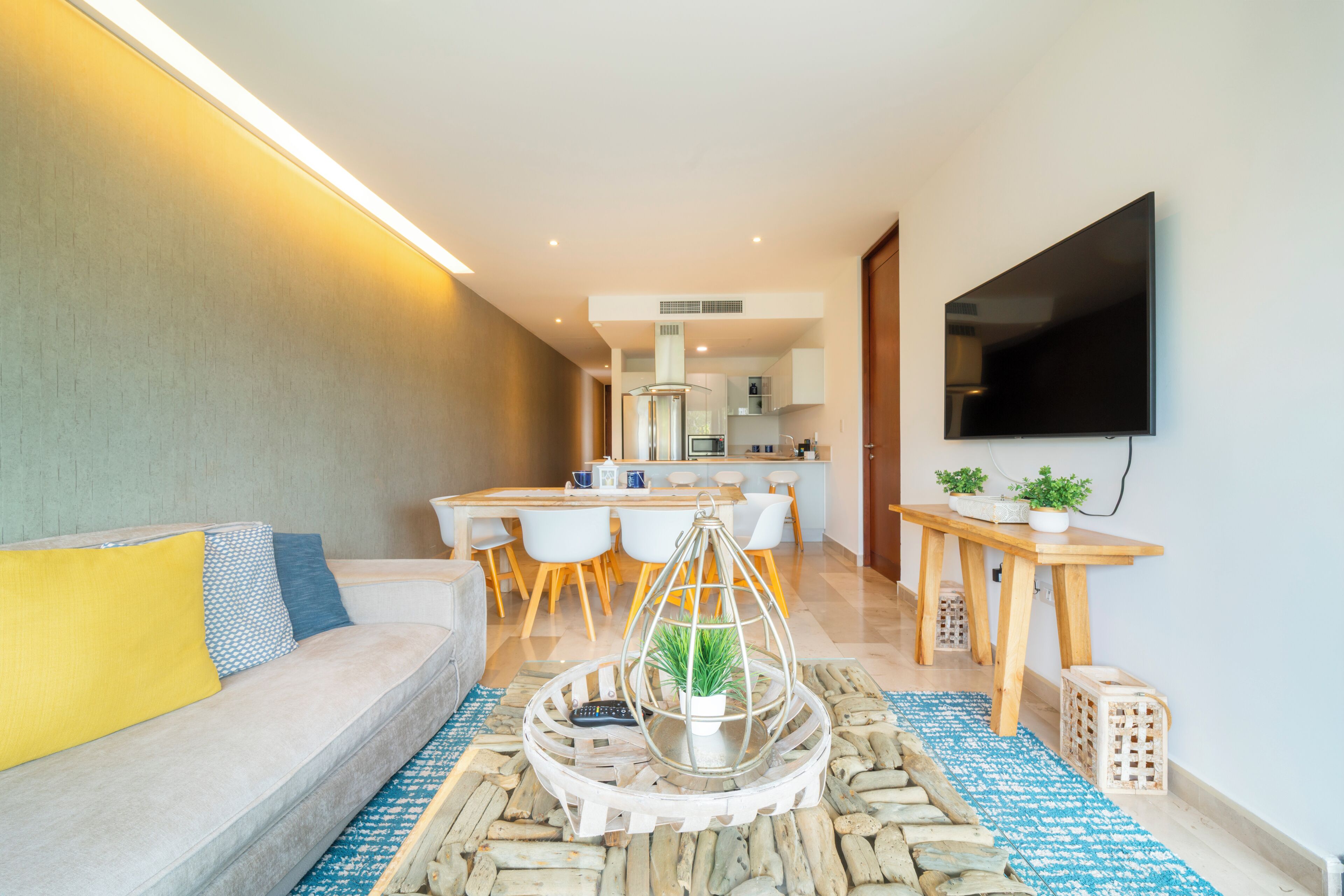 A modern living room with a sofa, a coffee table with a plant, a dining area with chairs, and a kitchen in the background.