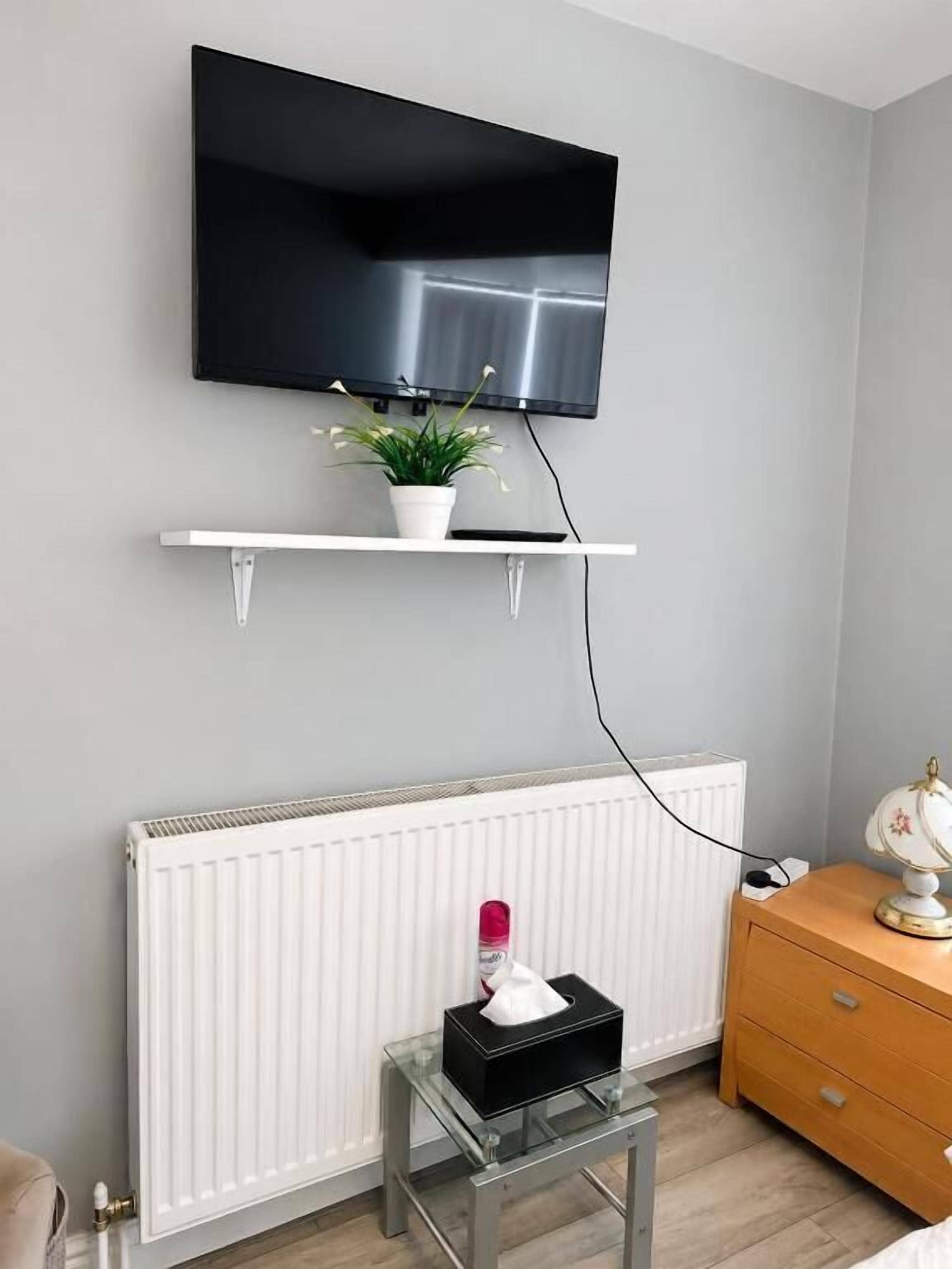 Huoneessa on seinälle asennettu televisio, valkoinen patteri, lasinen pöytä, jonka päällä on laatikko, sekä puinen lipasto.