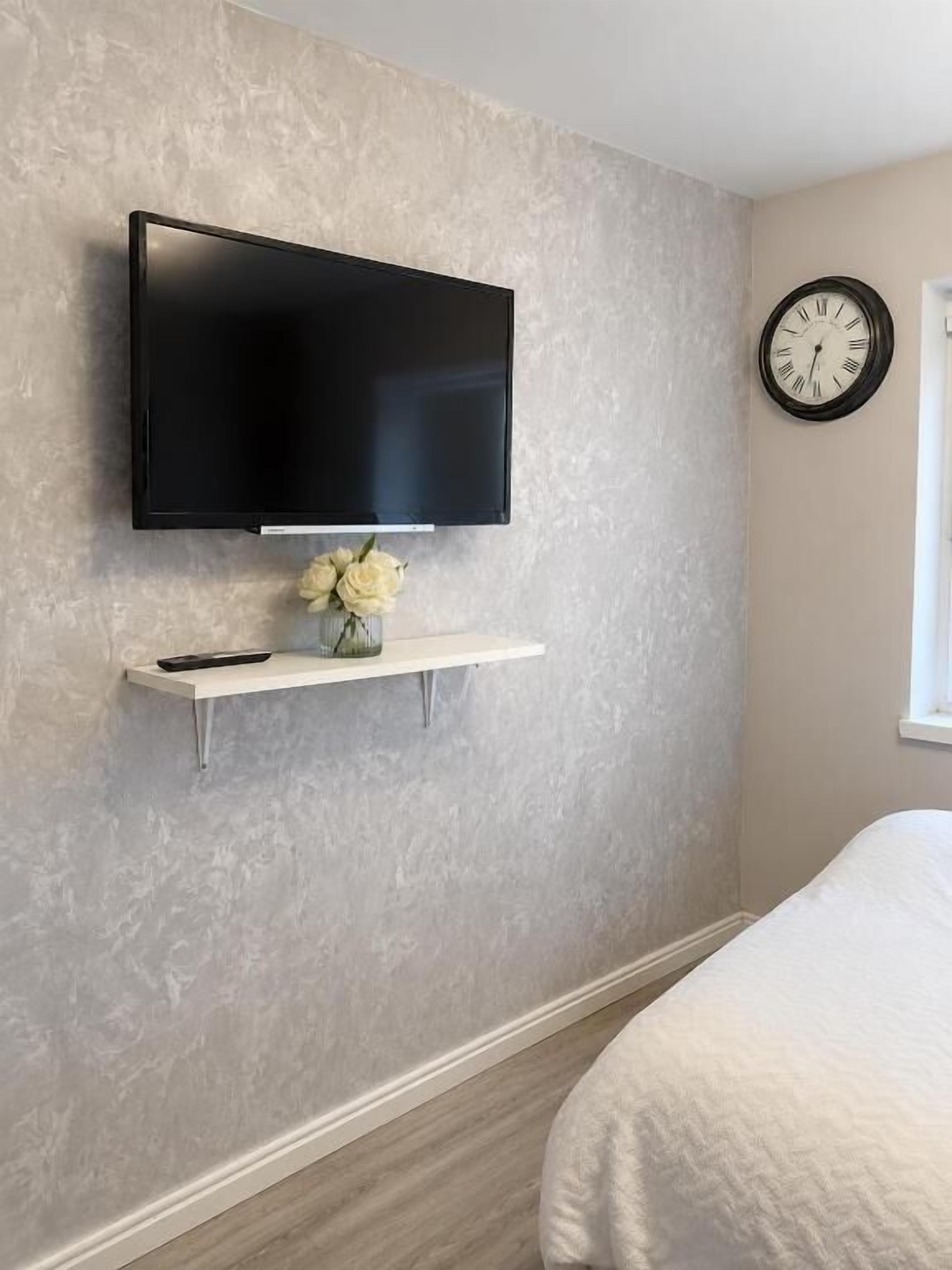 Une chambre à coucher avec un téléviseur fixé au mur, une horloge, une étagère avec un vase et un lit.