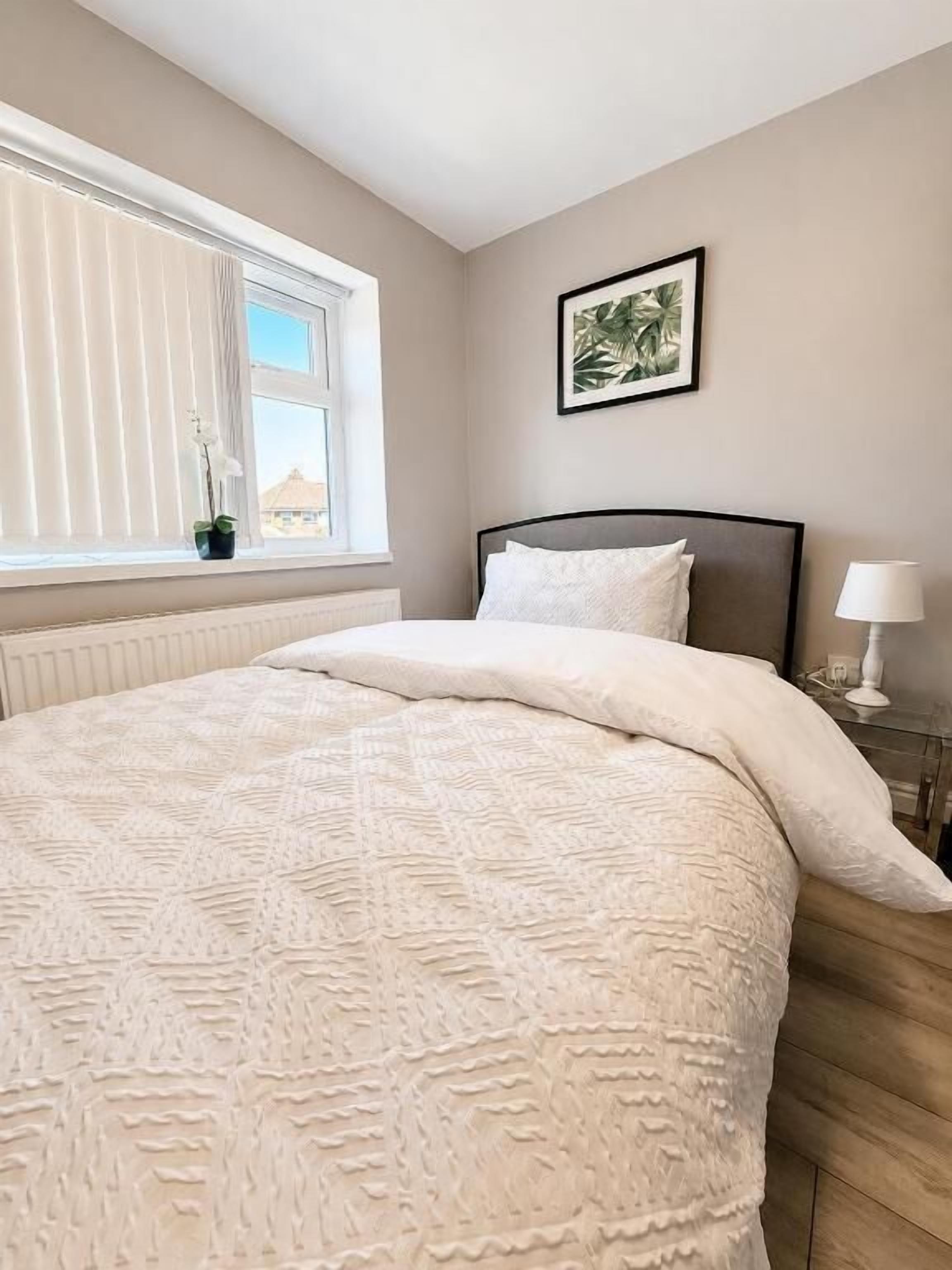 A bedroom with a bed, a window with blinds, a bedside table with a lamp, and a framed picture on the wall.