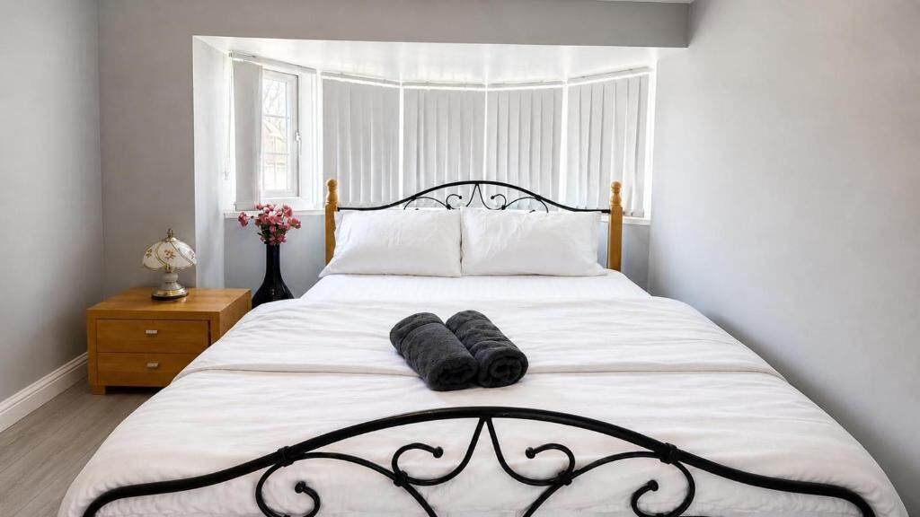 Una cama bien tendida con sábanas blancas y toallas enrolladas, una mesita de noche con lámpara y jarrón, y una ventana con cortinas blancas.