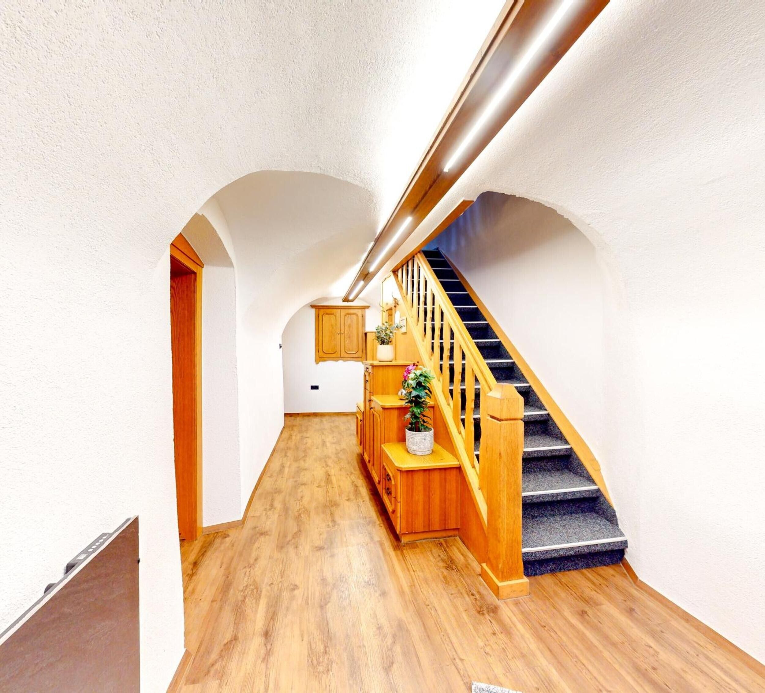 A hallway with wooden flooring, a staircase with wooden railings, and a wooden cabinet with a plant.