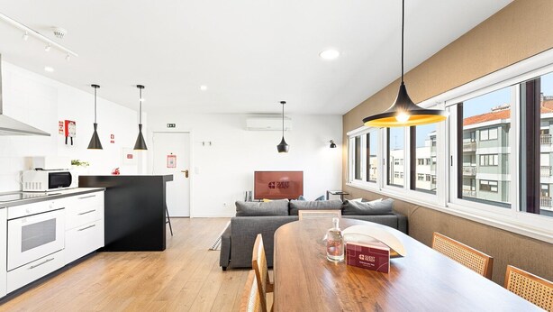 Une cuisine moderne avec des meubles blancs, un four à micro-ondes et un îlot central noir. On y trouve un coin repas avec une table et des chaises en bois, un canapé et un panneau indiquant « PRÊT POUR LES INVITÉS ».