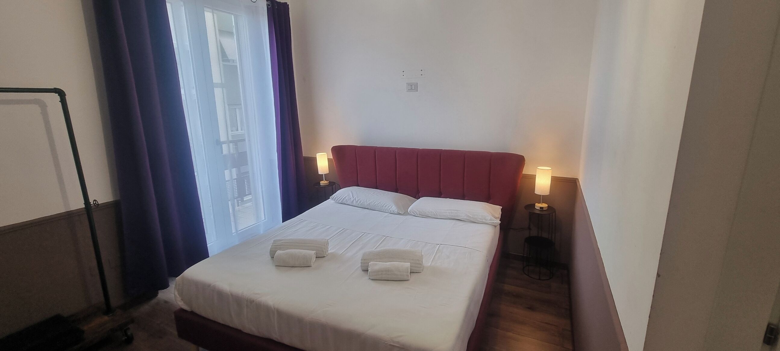 A hotel room with a bed, two bedside lamps, a window with curtains, and a wall socket.