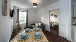 A modern living room with a dining table set for four, a grey sofa, a television, and a window with curtains. - Contractor Room • Kitchen • Central Spot (Neath)