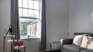 A living room with a grey sofa, a small table with a lamp, a radiator, and a window with curtains. - Contractor Room • Kitchen • Central Spot (Neath)