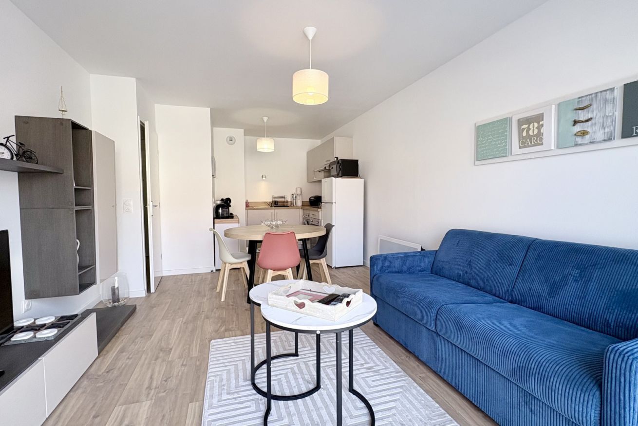 A modern living room with a blue sofa, a dining area with a table and chairs, and a kitchen in the background.