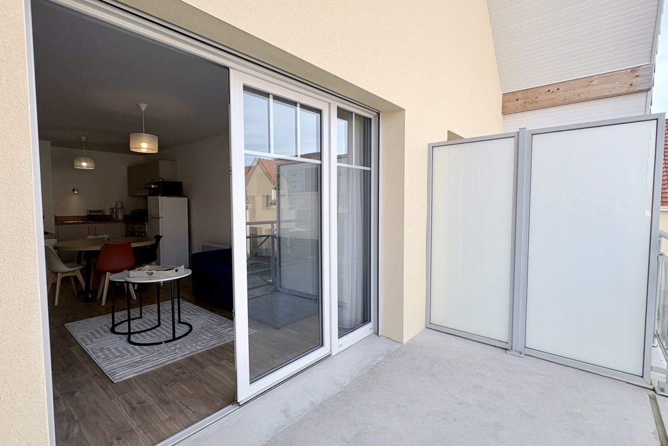 A modern apartment with a sliding glass door leading to a balcony, a dining area with a table and chairs, and a kitchen visible through the door.