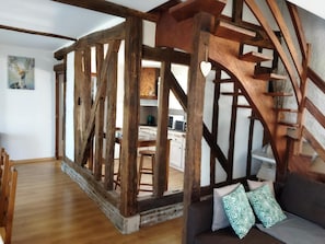 Ein rustikales Interieur mit freiliegenden Holzbalken, einer Holztreppe, einer Couch mit gemusterten Kissen und einer durch einen offenen Durchgang sichtbaren Küche.
