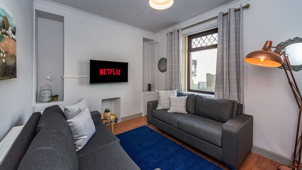 A modern living room with a grey sofa, a blue rug, a TV displaying 'NETFLIX,' a floor lamp, and a window with curtains. - 5 Bed Home • Parking • Ideal for Contractors (Carmarthenshire)