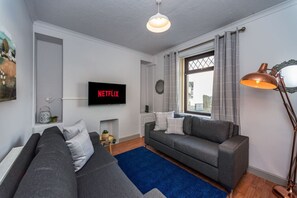 Ein modernes Wohnzimmer mit einem grauen Sofa, einem blauen Teppich, einem Fernseher, der 'NETFLIX' anzeigt, einer Stehlampe und einem Fenster mit Vorhängen.