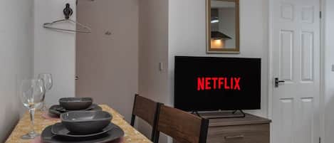 Un comedor con una mesa dispuesta para dos personas, un televisor mostrando 'NETFLIX', un espejo y una puerta.