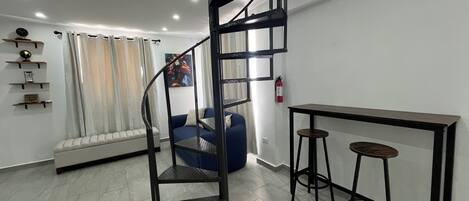 Een moderne leefruimte met een spiltrap, een blauwe bank, een klein tafeltje met krukken en een brandblusser aan de muur.