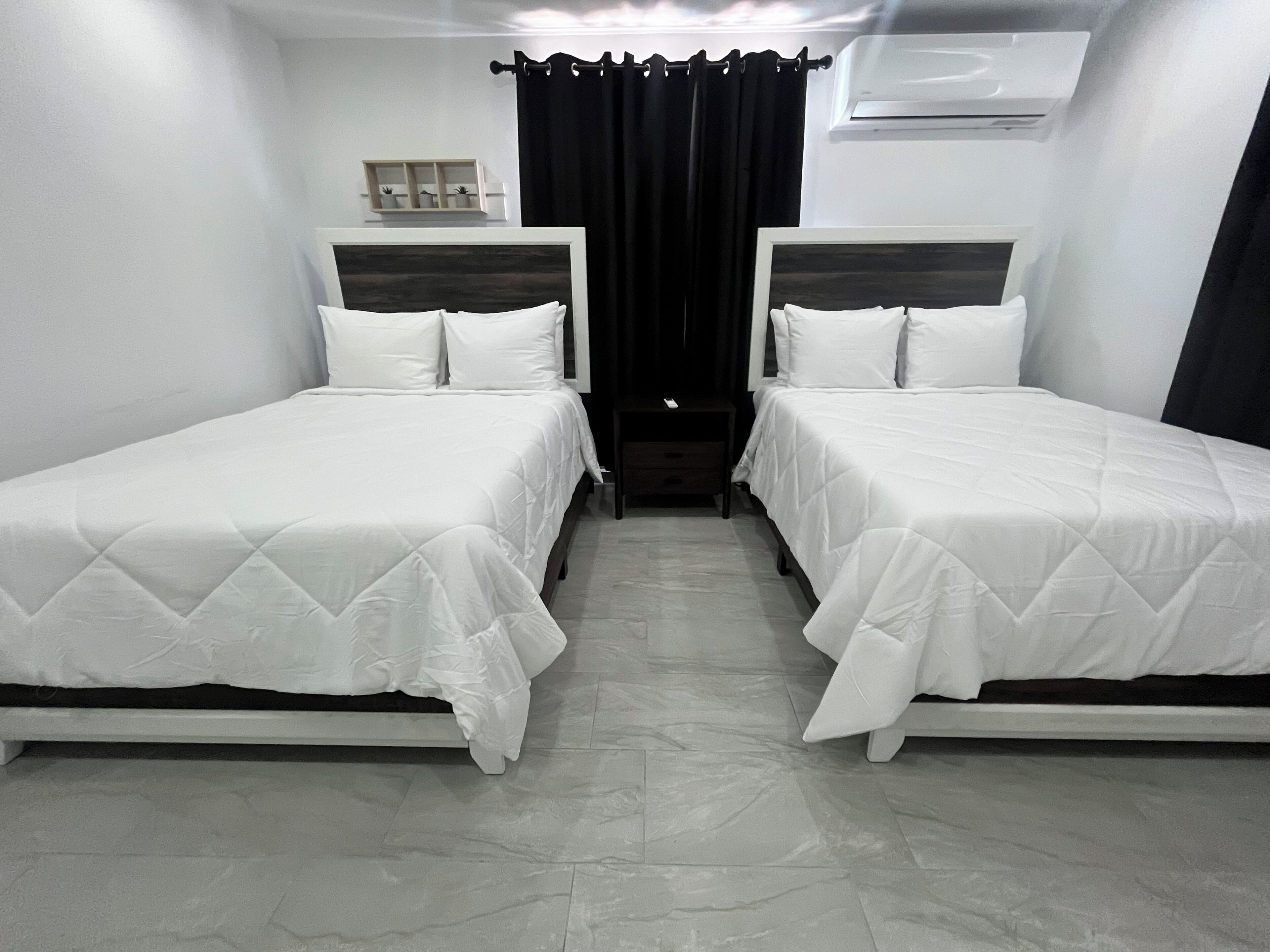 Une chambre d’hôtel avec deux lits simples, une petite table de chevet, une étagère avec des objets décoratifs et une fenêtre avec des rideaux.