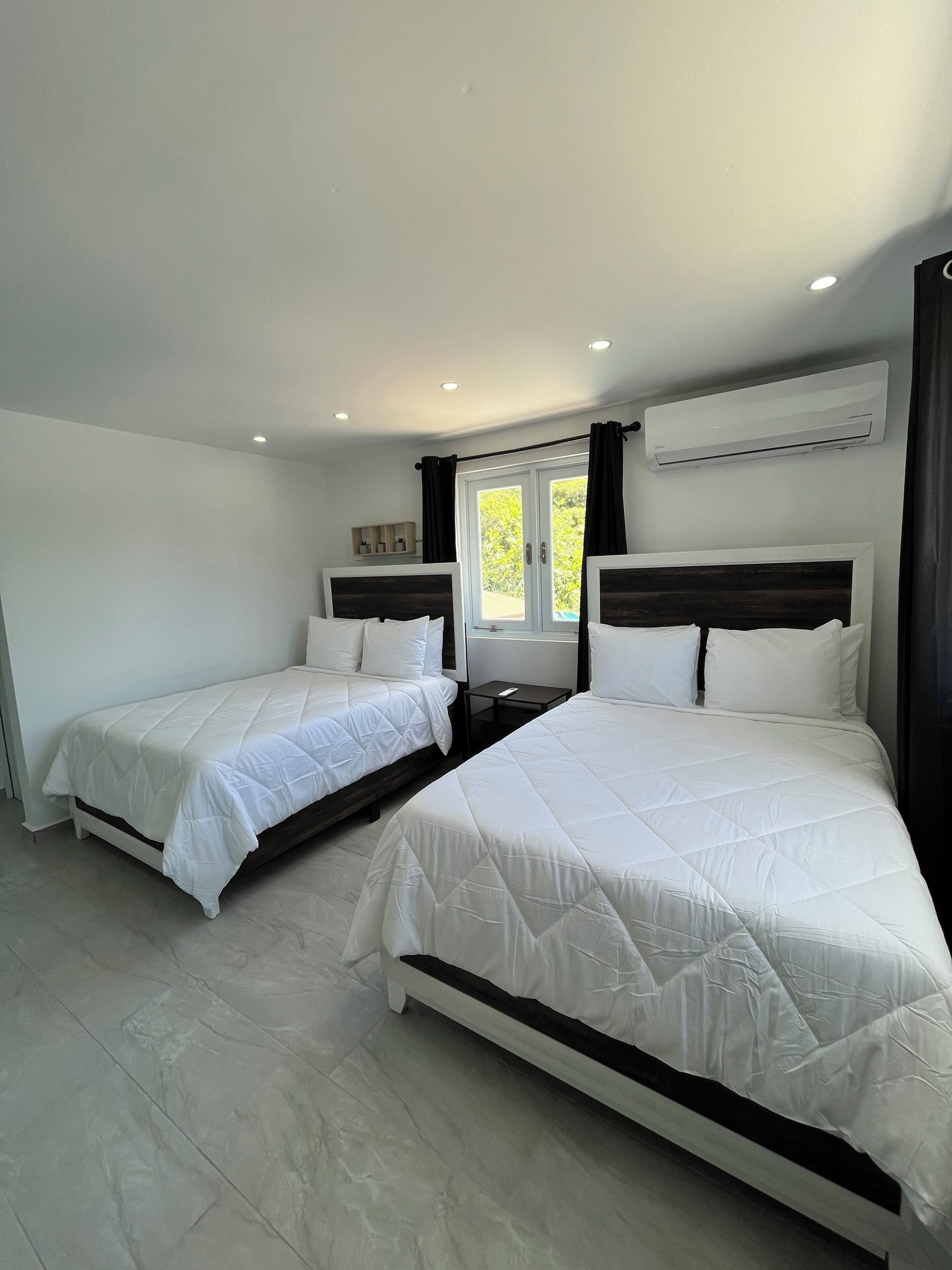 A hotel room with two beds, a window with curtains, an air conditioning unit, and a small bedside table.