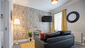 A modern living room with a stone wall, a fireplace, a black leather sofa, a mounted TV, and a yellow rug. - Spacious Home • Parking • Kitchen • Ideal for Work (Port Tennant)