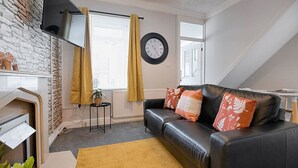 A modern living room with a black leather sofa, a yellow rug, a fireplace, a clock, and a window with curtains. - Spacious Home • Parking • Kitchen • Ideal for Work (Port Tennant)