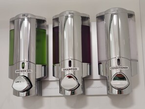 Three chrome dispensers with labels for shower gel, shampoo, and conditioner.