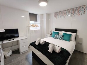 A modern hotel room with a bed, a desk with a TV, a small window, and a wall-mounted decoration.