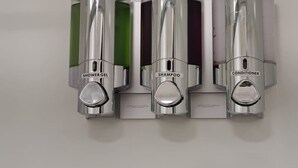 Drei metallene Spender mit den Aufschriften Duschgel, Shampoo und Conditioner an einer Wand.