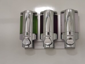 Drei metallene Spender mit den Aufschriften Duschgel, Shampoo und Conditioner an einer Wand.