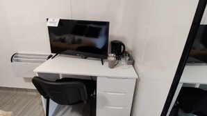 Ein weißer Schreibtisch mit einem schwarzen Monitor, einem schwarzen Bürostuhl, einer Kaffeemaschine und einem weißen Heizkörper.