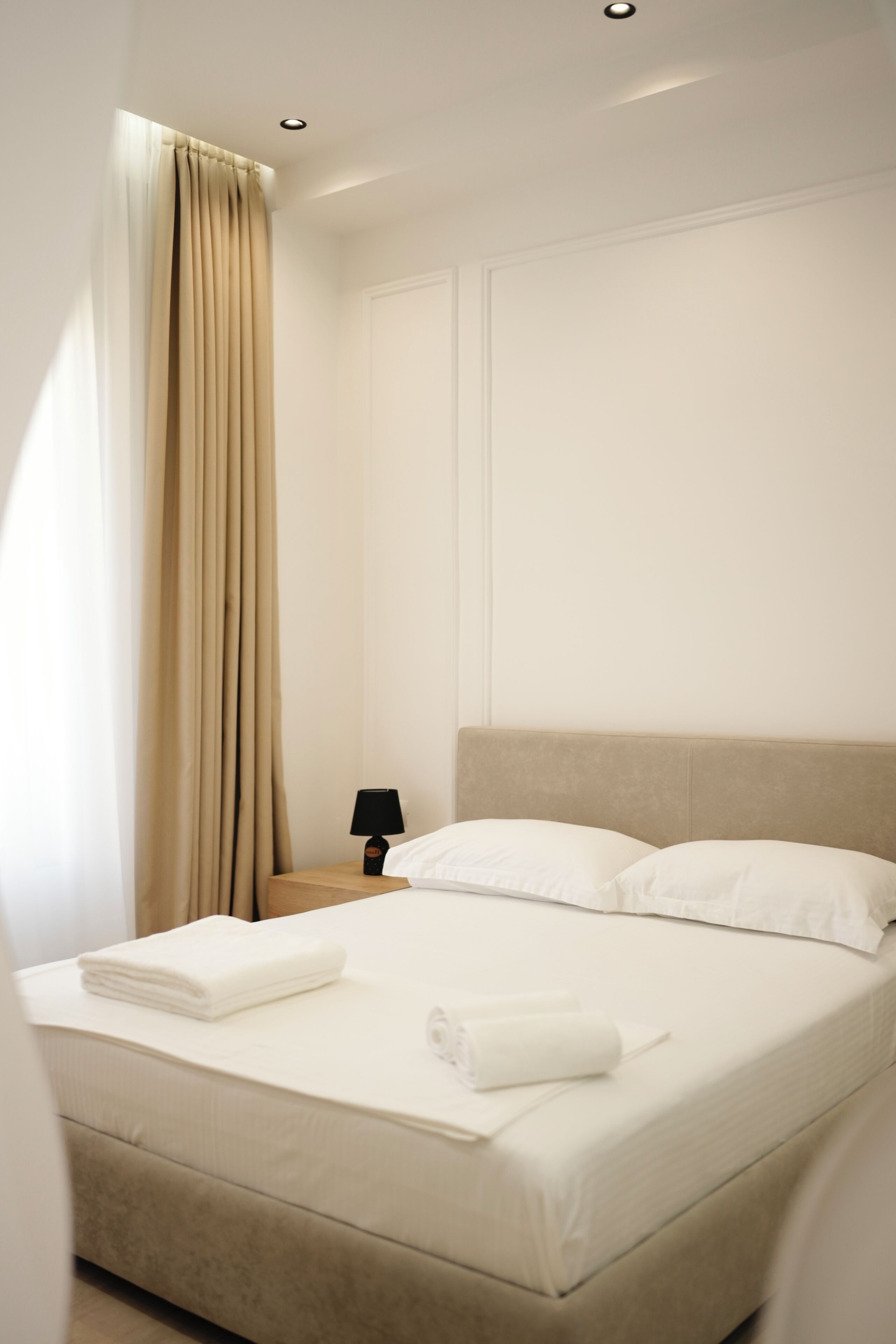A neatly made bed with white linens, a bedside lamp, and a window with beige curtains.