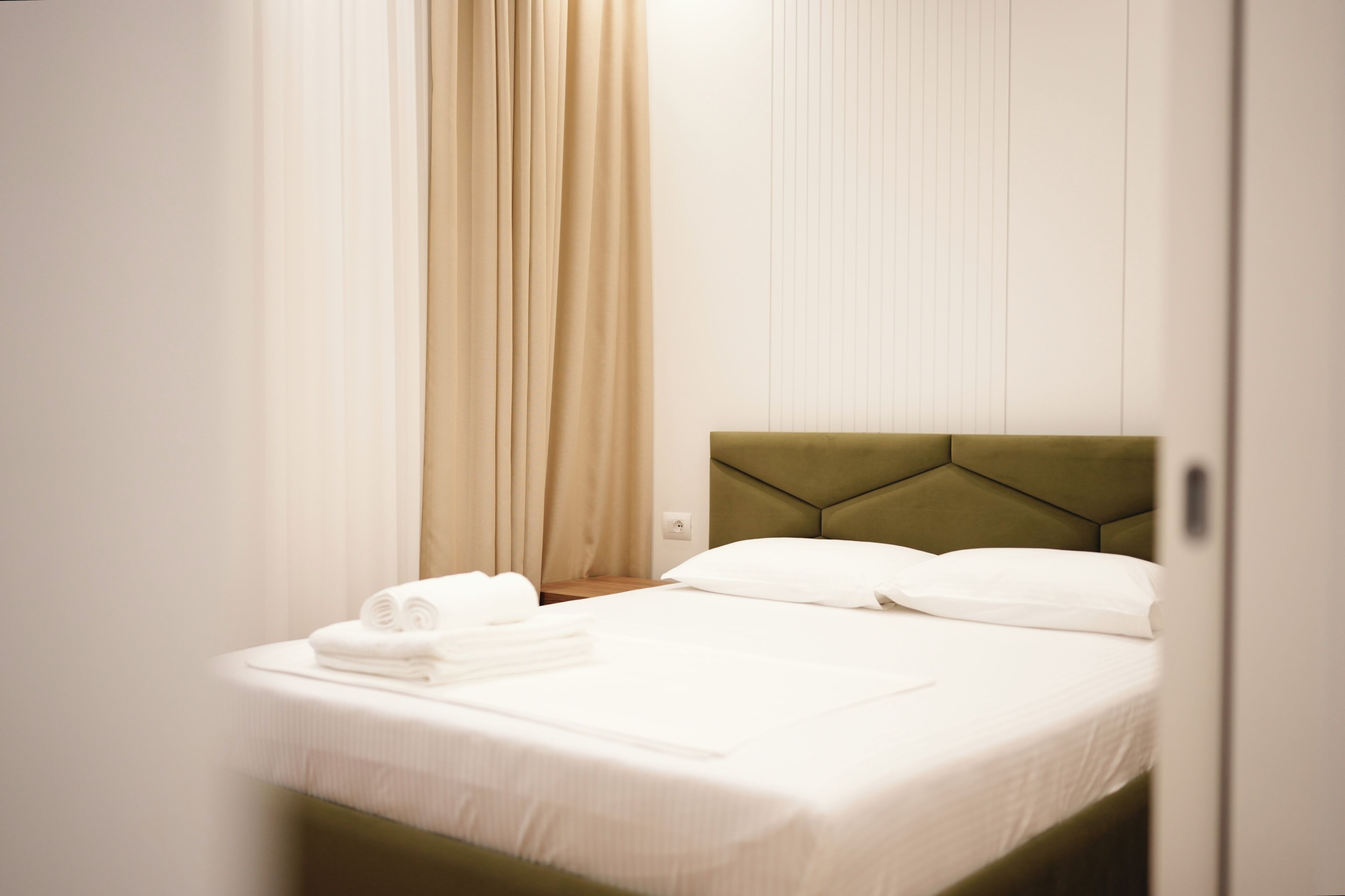 A neatly made bed with white linens, a green upholstered headboard, and a set of folded white towels on the side.