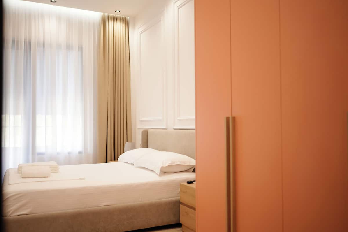 A neatly made bed with white linens, a wooden nightstand, and a wardrobe with a light wood finish.