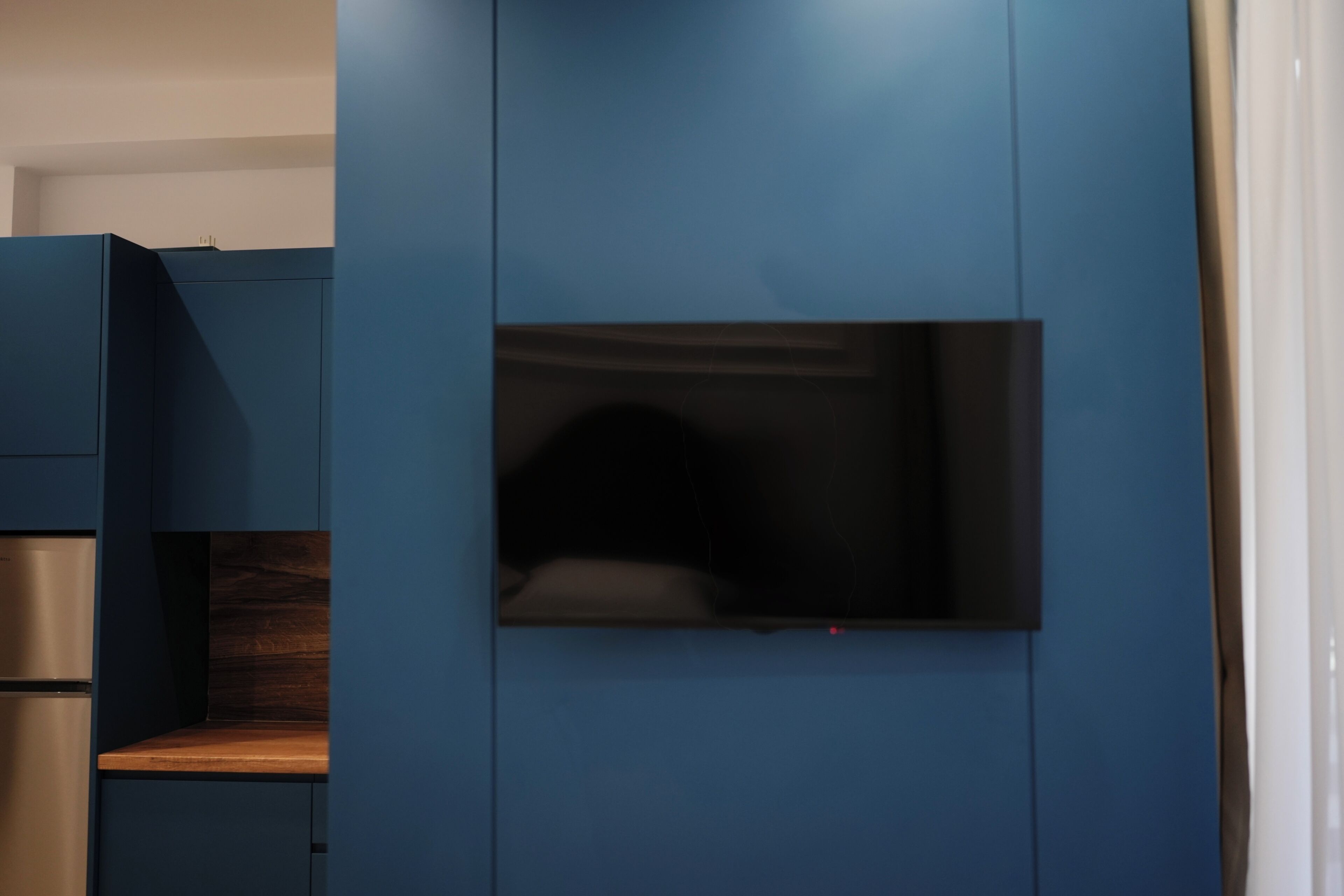 A modern kitchen with blue cabinetry, a built-in oven, and a flat-screen TV mounted on the wall.