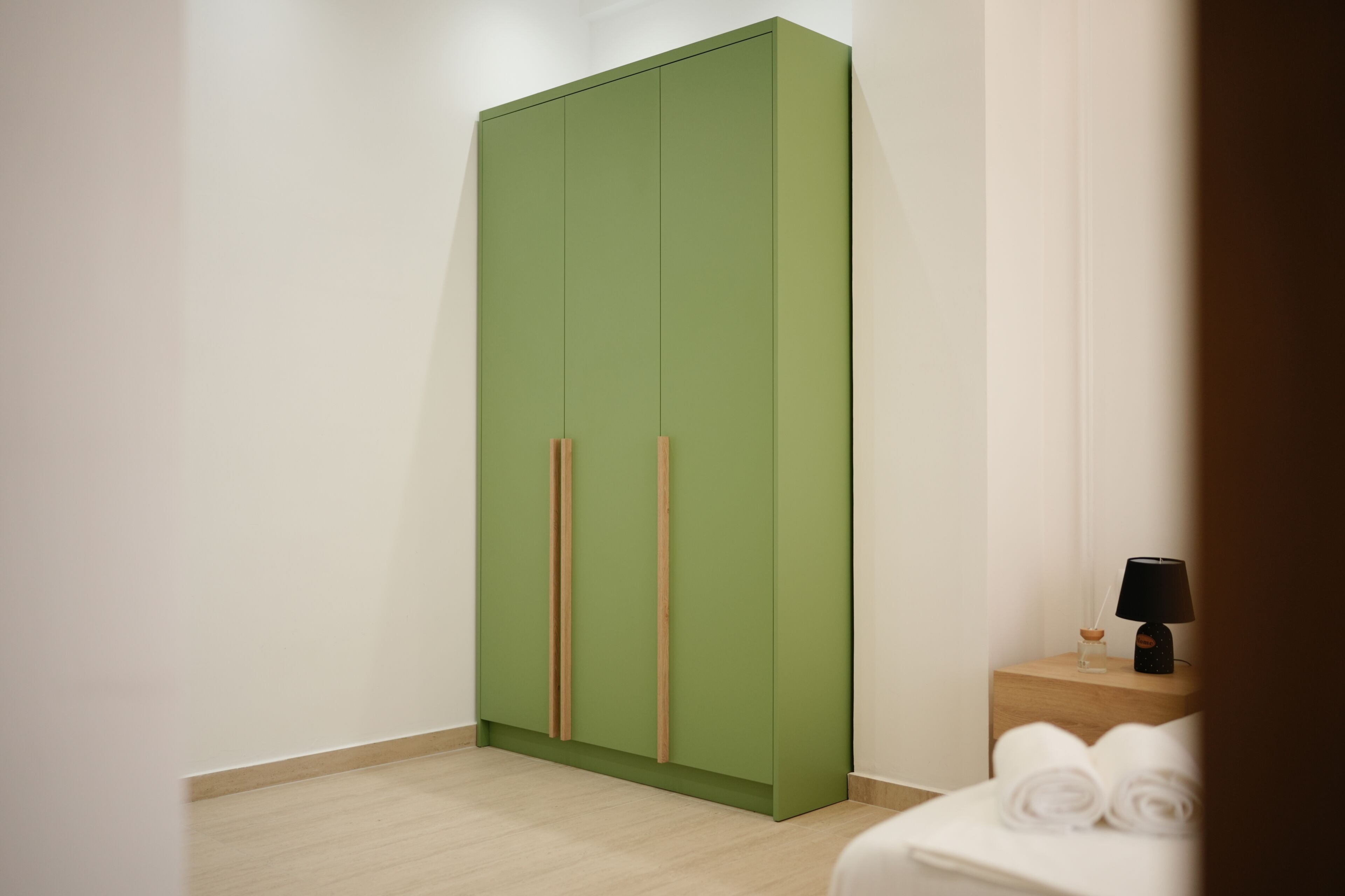 A room with a green wardrobe, a wooden nightstand with a lamp, and a bed with rolled towels.