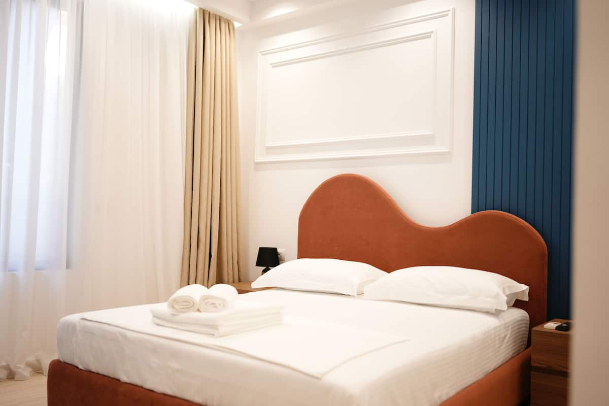 A neatly made bed with white linens and a brown headboard, a nightstand with a lamp, and a window with sheer curtains.