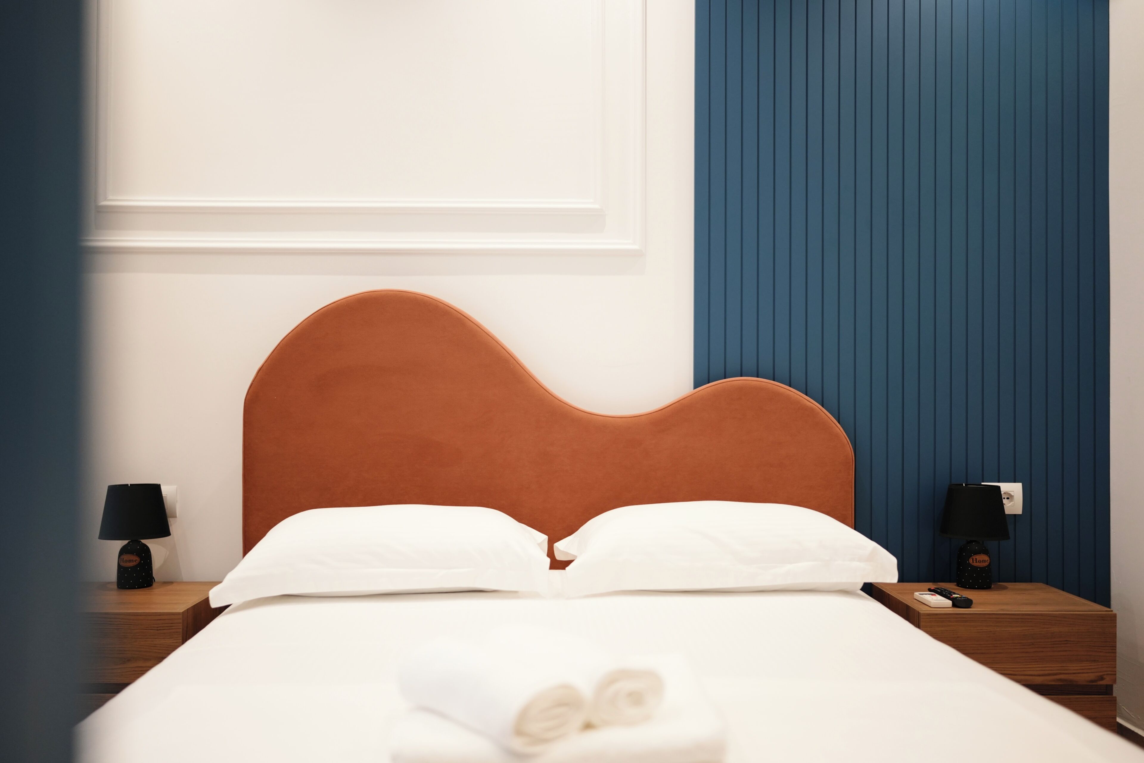 Una cama bien hecha con sábanas blancas, un cabecero marrón y una toalla doblada al pie. Hay dos mesitas de noche con lámparas y un interruptor de luz en la pared.