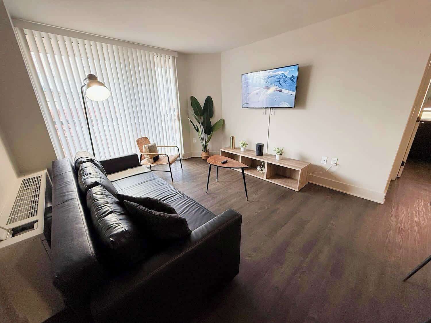 A living room with a black leather sofa, a wooden coffee table, a TV mounted on the wall, and a large window with blinds.