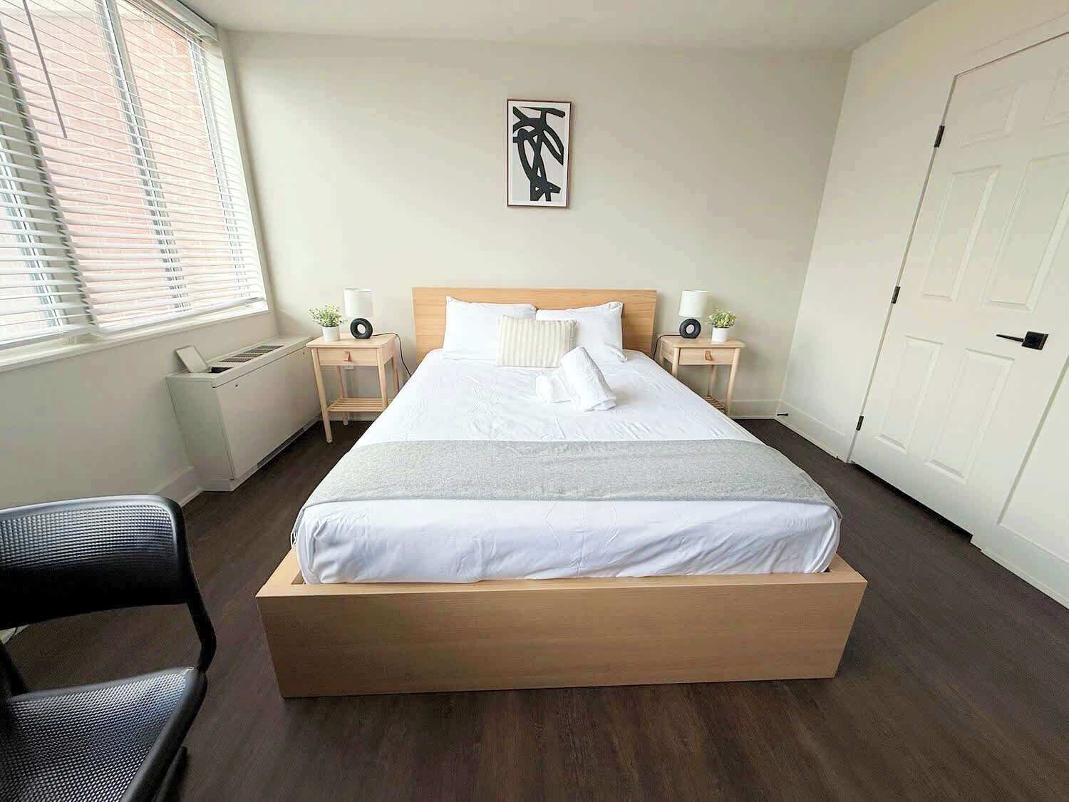 A hotel room with a bed, bedside tables, a chair, a window with blinds, a door, and a wall-mounted artwork.
