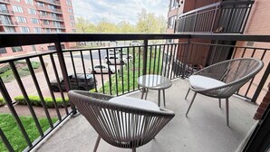 En balkon med fletstole og et rundt bord, med udsigt over en parkeringsplads og bygninger.