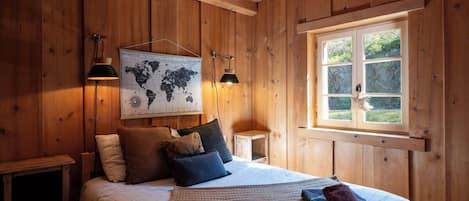 Una habitación con paneles de madera, una cama, dos lámparas fijadas a la pared, un mapa mundial y un ventanal con vistas a zonas verdes.